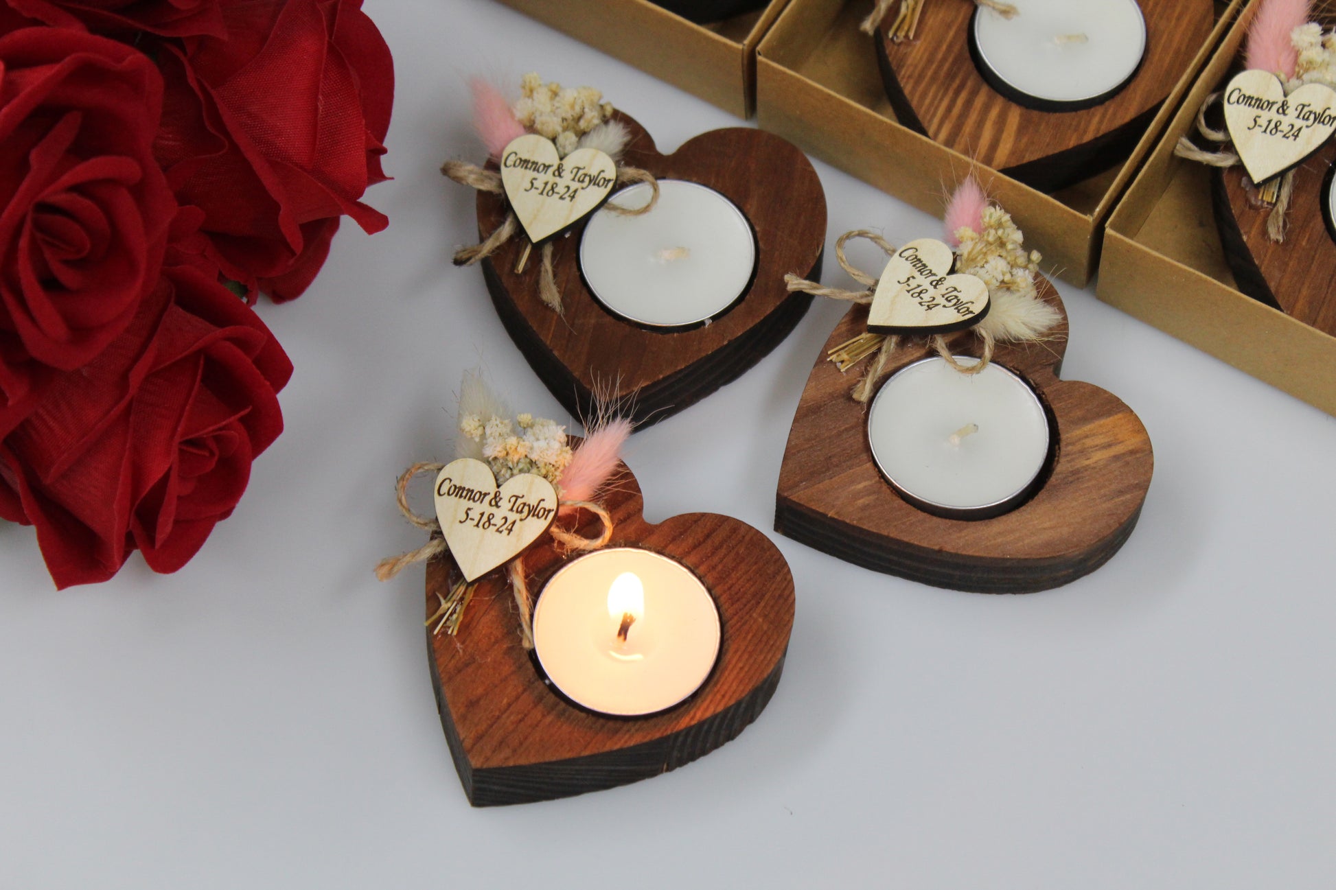 Charming Personalized Heart-Shaped Wooden Candle Holders with Dried Flowers. These elegant wedding favors feature a rustic wooden heart design, a burning tealight candle, and a personalized wooden heart tag with delicate dried flower arrangements. Perfect for adding a unique and romantic touch to any wedding celebration. Example names and dates are shown for personalization.