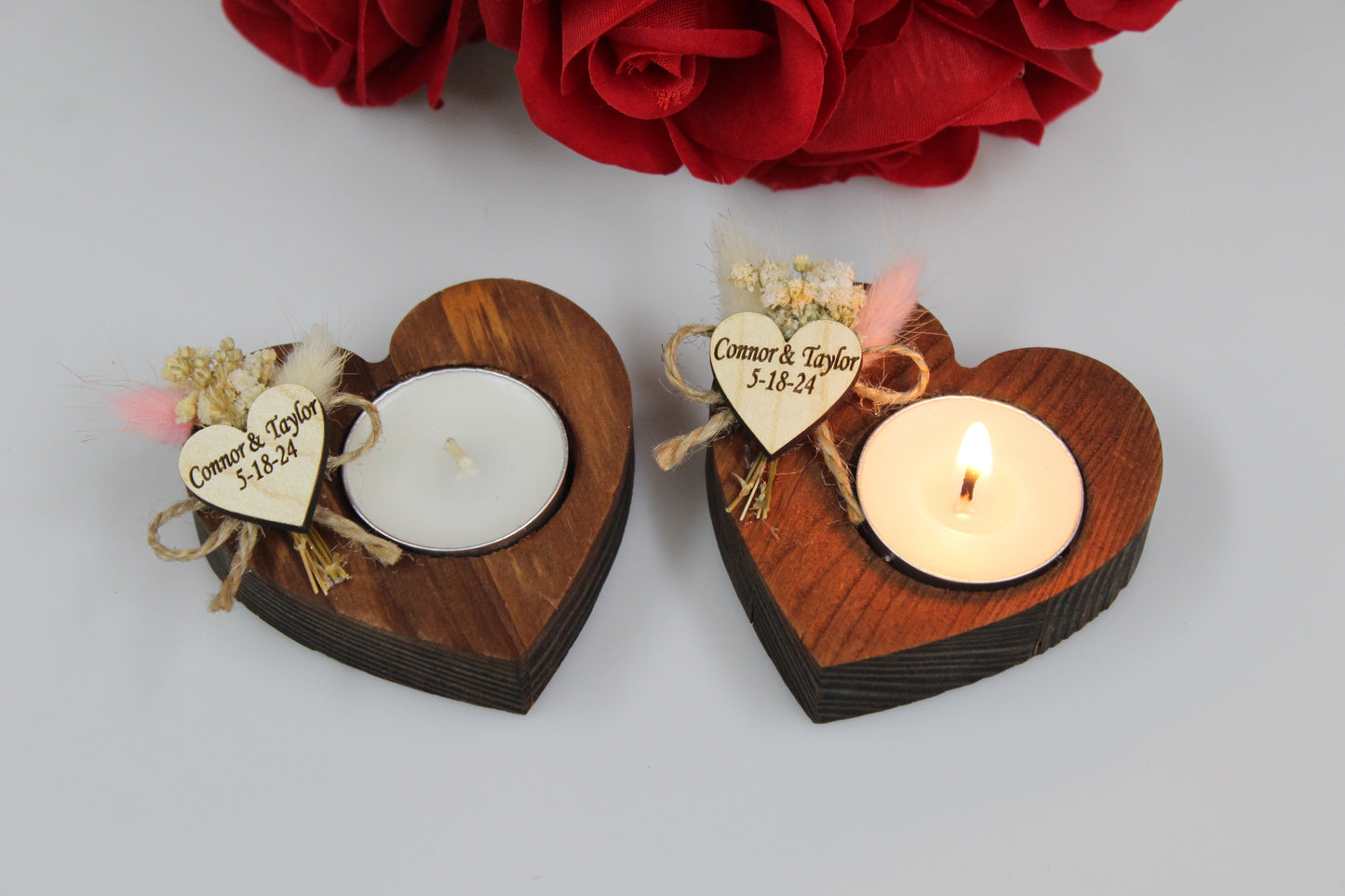 Personalized Heart-Shaped Wooden Candle Holders with Dried Flowers. These exquisite wedding favors feature rustic wooden heart candle holders, one with a burning tealight candle and the other unlit, adorned with a personalized wooden heart tag and delicate dried flower arrangements. Ideal for adding a unique and romantic touch to any wedding celebration. Example names and dates are shown for personalization.