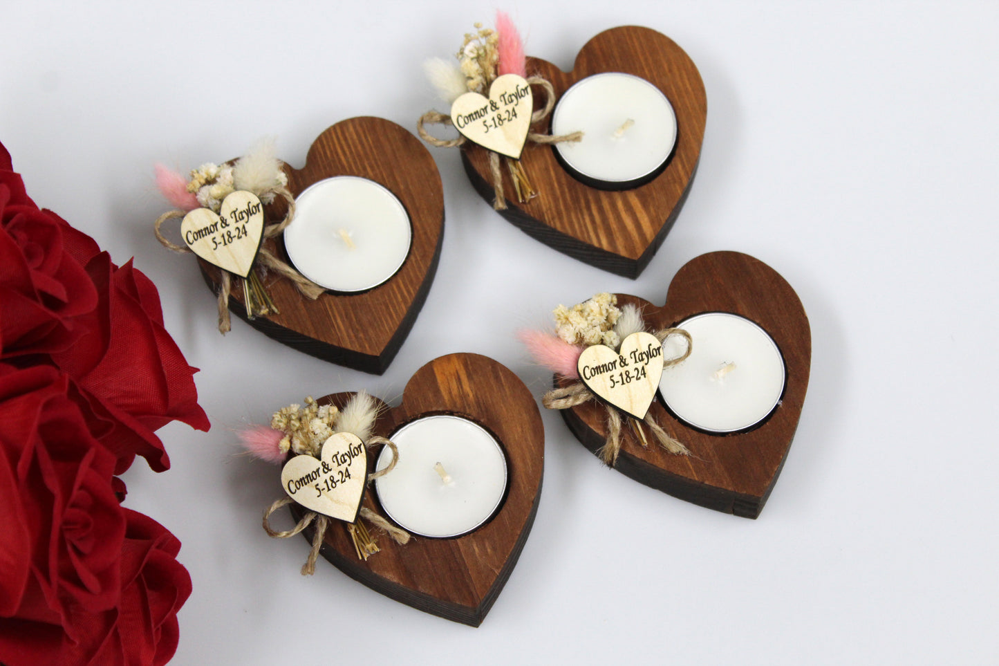 Elegant and personalized heart-shaped wooden candle holders adorned with dried flowers, ideal for wedding favors. Each candle holder features a tea light and a customizable wooden heart tag with names 'Connor & Taylor' and date '5-18-24' as examples, showcasing the personalized touch. These rustic, handcrafted wedding favors are perfect for adding a sentimental and unique element to wedding celebrations. Displayed with red roses, highlighting their romantic appeal.