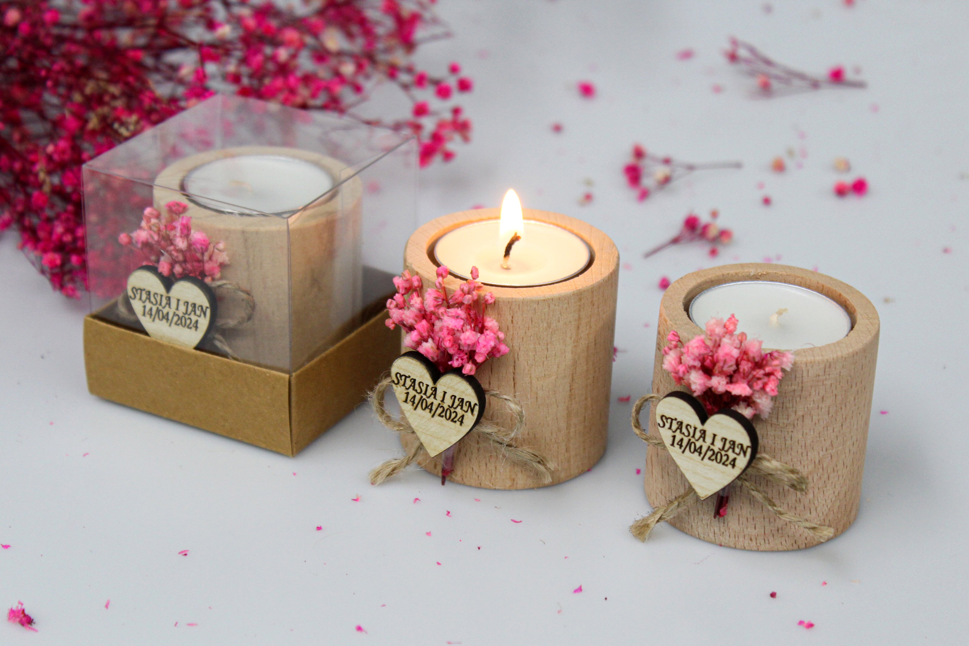 Personalized wooden tea light candle holders, showcasing custom names and dates as examples. Each holder features pink dried flowers and a heart-shaped tag engraved with 'Stasia & Jan 14/04/2024'. One candle holder is boxed, one is lit, and one is unlit, illustrating the personalized nature of these wedding favors. The background includes pink floral accents, enhancing the romantic theme.