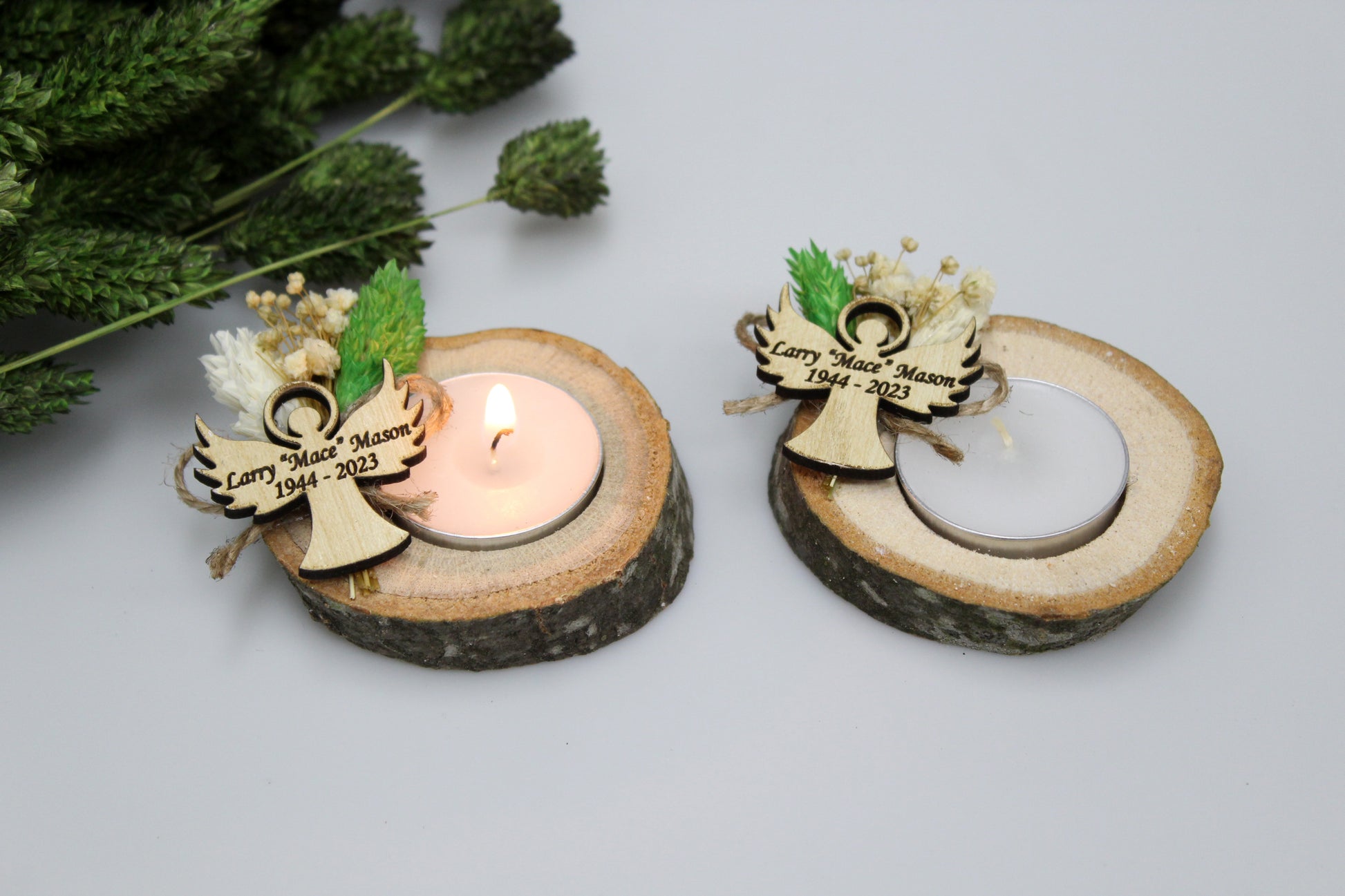 Personalized handcrafted rustic memorial candle holders with engraved angel tags. The image showcases two candle holders made from natural wood slices, each featuring a tealight candle and adorned with dried flowers. One candle is lit, and both have custom engraved angel tags with the name 'Larry "Mace" Mason' and dates '1944-2023'. Perfect for personalized funeral favors, these memorial candle holders offer a heartfelt tribute.