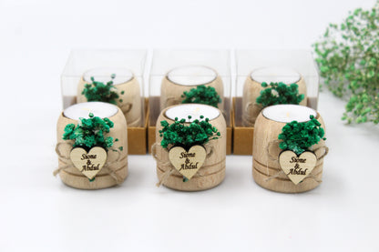 Rustic wedding display featuring six personalized wooden candle holders adorned with green dried flowers and custom engraved heart-shaped tags, with names and dates as examples. The handcrafted tealight holders are elegantly presented in a combination of boxed and unboxed arrangements, emphasizing their bespoke charm and attention to detail. Ideal for adding a personalized touch to wedding decor or as memorable keepsakes for guests.