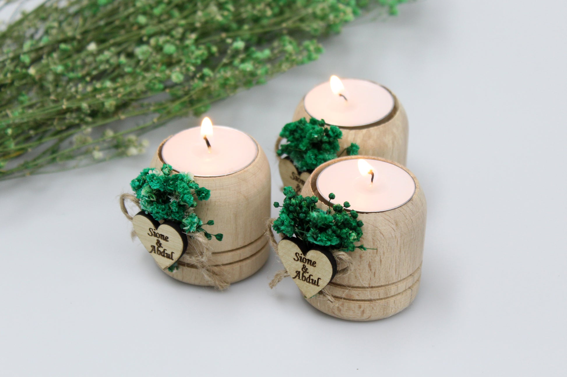 Three personalized wooden candle holders adorned with green dried flowers and custom engraved heart tags featuring the names "Simone & Akbul" as examples for users. Perfect for adding a rustic charm and romantic ambiance to weddings and special celebrations.