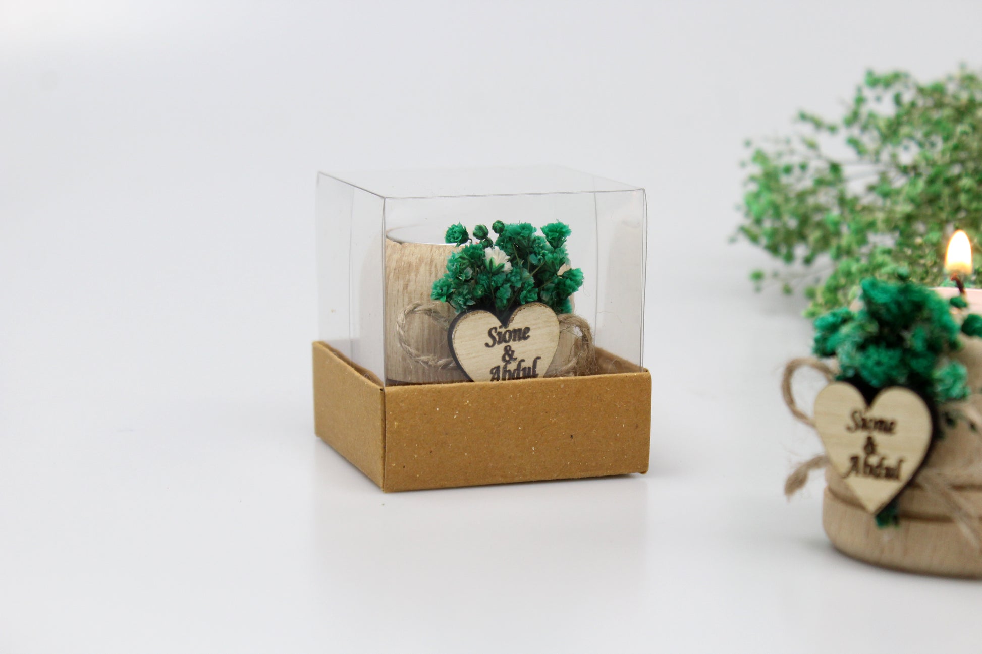 Handcrafted wooden candle holders adorned with preserved green flowers and personalized heart-shaped tags featuring names and dates as examples. Each favor is elegantly packaged in a clear box, perfect for adding a touch of rustic charm to weddings or special occasions.