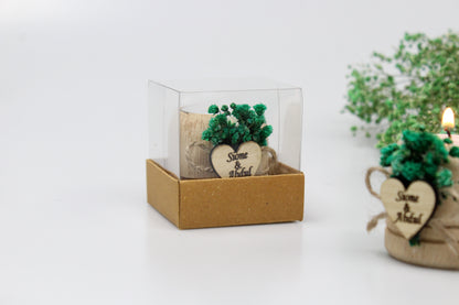 Handcrafted wooden candle holders adorned with preserved green flowers and personalized heart-shaped tags featuring names and dates as examples. Each favor is elegantly packaged in a clear box, perfect for adding a touch of rustic charm to weddings or special occasions.