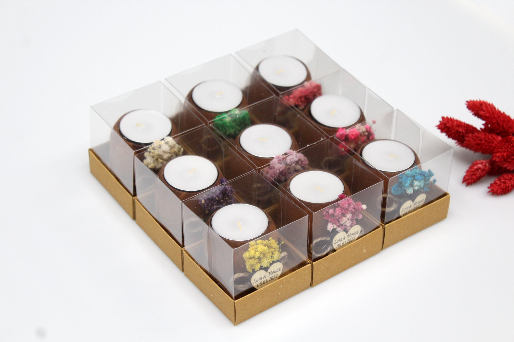 Set of nine handcrafted wooden tea light candle holders, each adorned with different colors of dried flowers and personalized heart-shaped tags. Packaged in clear boxes, these rustic wedding favors feature example names and dates, showcasing their customizable nature.