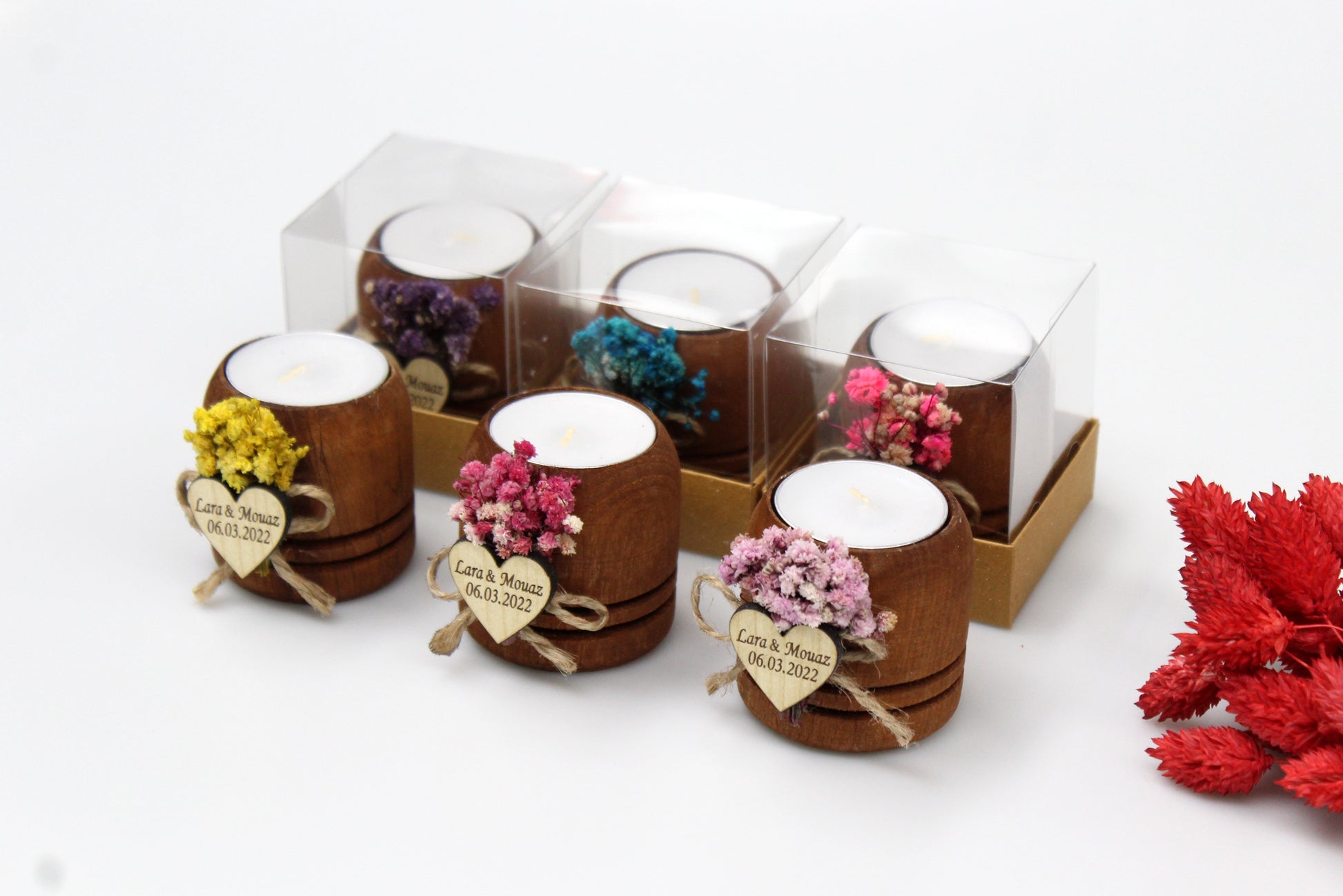 Rustic wooden tea light candle holders with personalized wooden heart tags displaying example names 'Lara & Mouaz' and date '06.03.2022'. Each holder is adorned with colorful dried flowers and packaged in individual clear boxes. These elegant wedding favors showcase various vibrant floral arrangements, adding a personalized touch to wedding decorations.