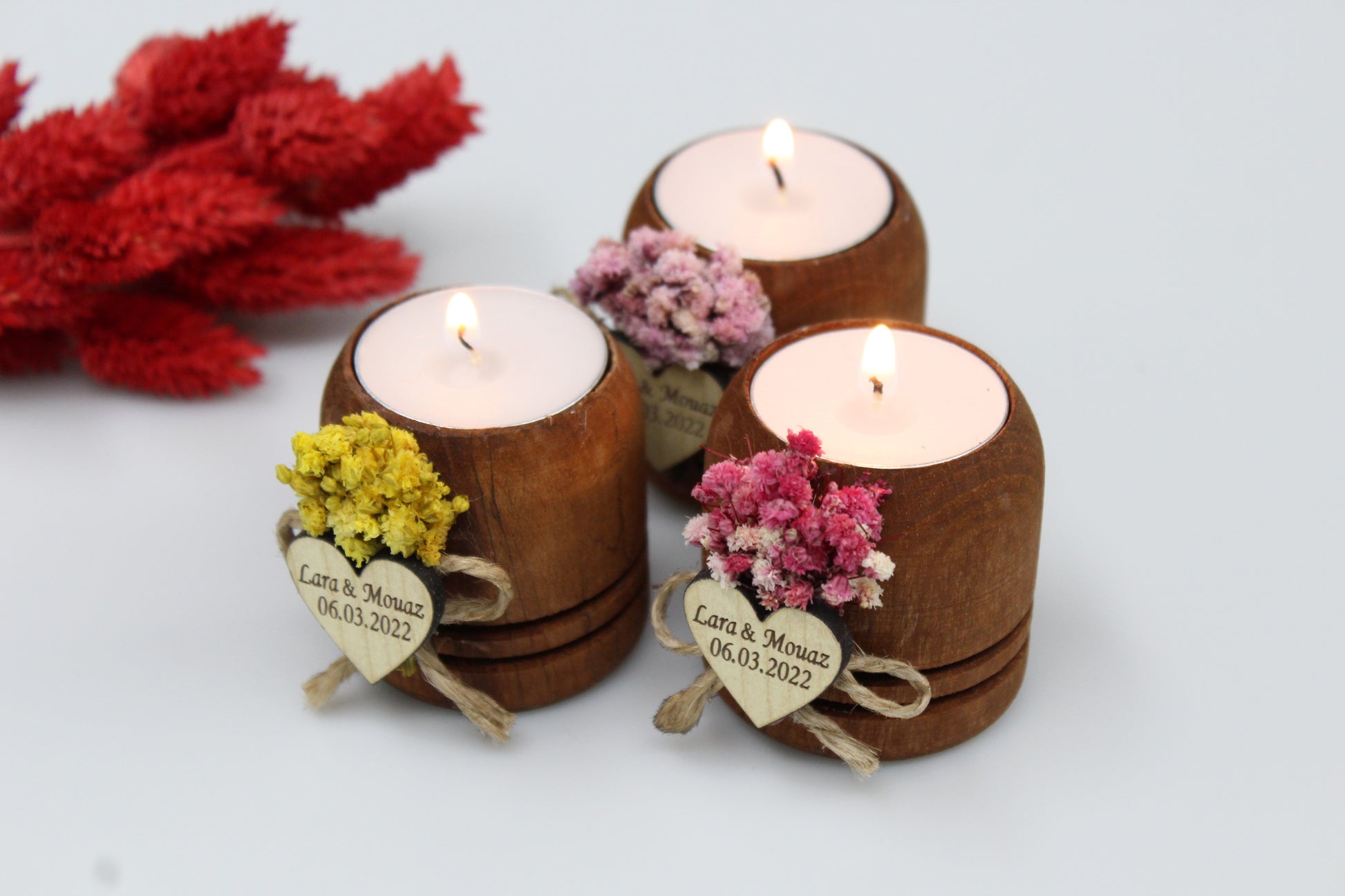 Rustic wooden tea light candle holders adorned with personalized wooden heart tags and colorful dried flower arrangements in yellow, pink, and red. Each holder is engraved with the example names 'Lara & Mouaz' and the date '06.03.2022'. The candles are lit, creating a warm and inviting ambiance, ideal for wedding favors.