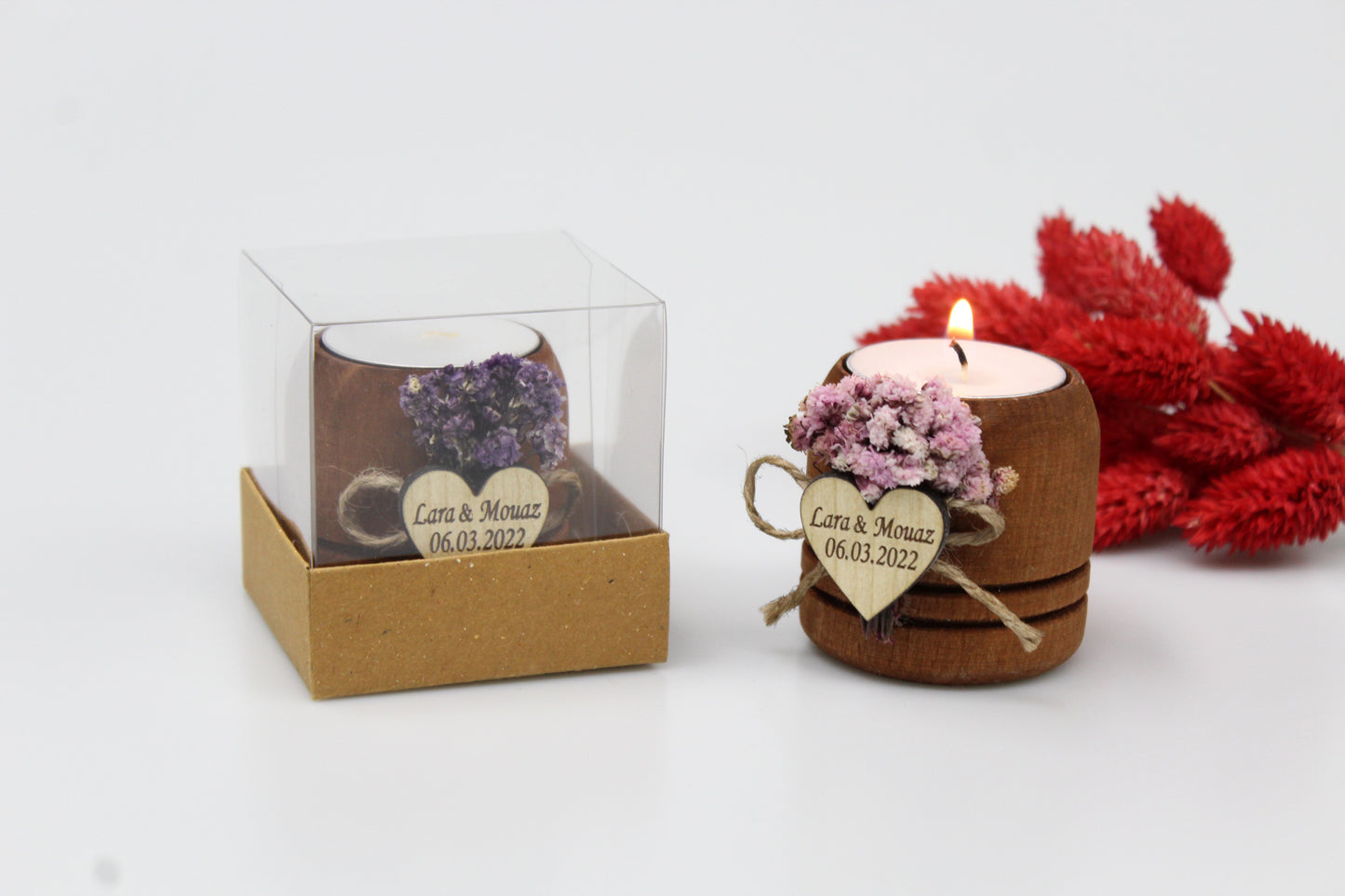 Personalized rustic wooden candle holders with dried flowers and custom heart-shaped tags. Example shown with names Lara & Mouaz and date 06.03.2022. One candle holder is lit, and the other is packaged in a clear box. Ideal for wedding favors, presented with red dried floral accents.