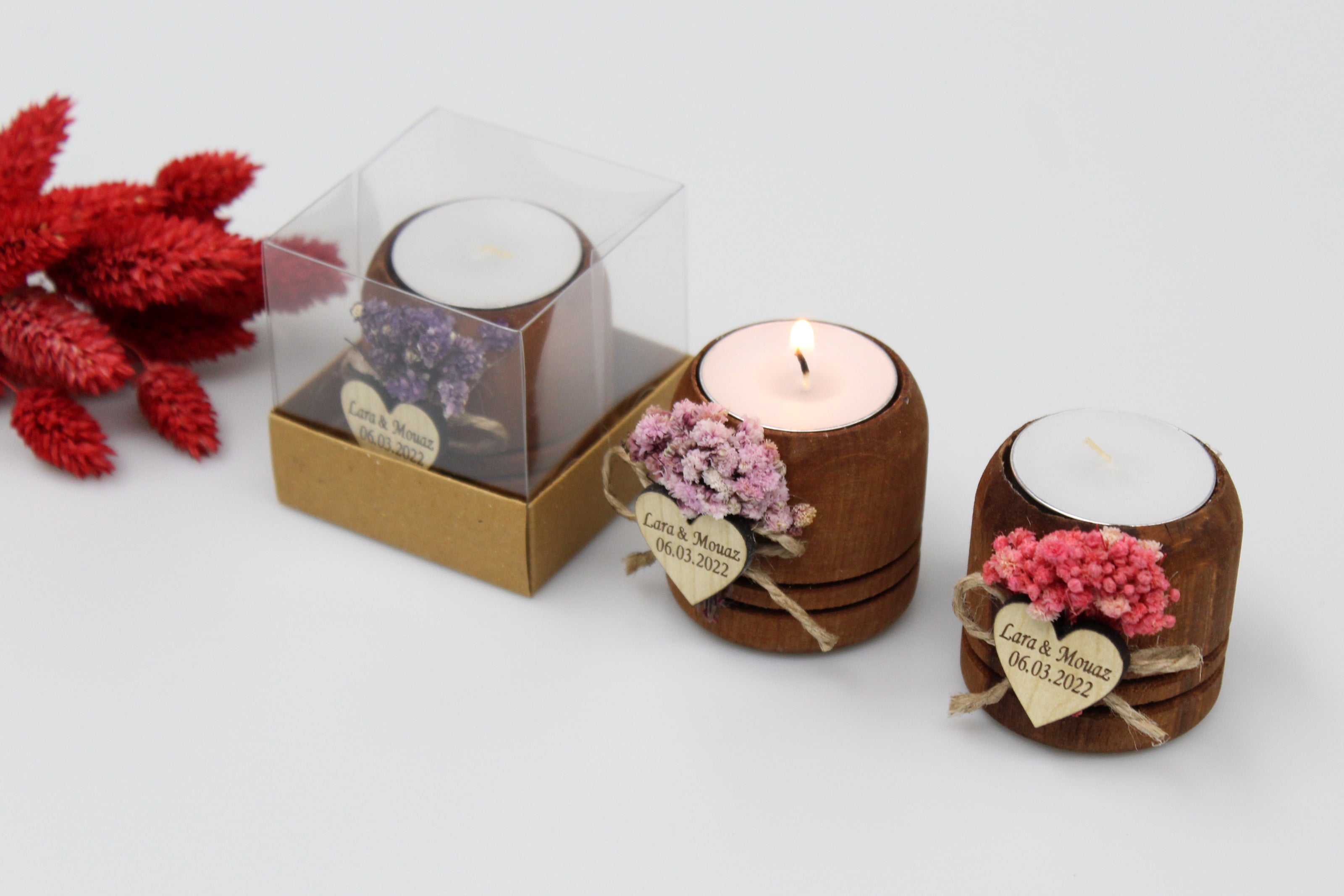 Handcrafted wooden tea light candle holders adorned with colorful dried flowers and personalized heart-shaped tags. One candle is packaged in a clear box, while two others are displayed lit and unlit. Ideal for rustic wedding favors, with example names and dates shown on the tags.