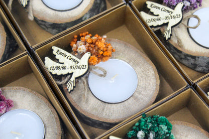 Close-up of personalized handcrafted rustic memorial candle holders featuring elegant dove-shaped tags engraved with the name "Karen Carney" and dates "12/09/60 - 05/16/23." These exquisite memorial favors rest on natural wood log bases, each beautifully adorned with vibrant dried orange flowers. Perfect for creating a heartfelt tribute to loved ones with sophistication and timeless elegance. Names and dates are examples to illustrate customization.