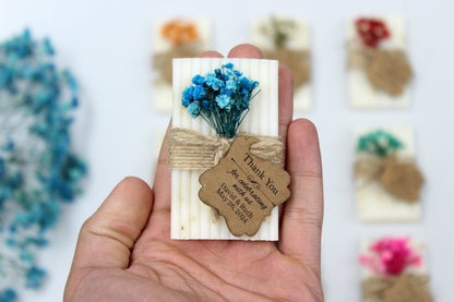 Handheld personalized handmade wedding soap favor adorned with blue dried flowers and rustic twine, featuring a custom thank you tag. Celebrate your special day with these elegant and eco-friendly wedding favors that delight guests with their charm and personal touch. Perfect for weddings, these handcrafted soaps offer a unique and memorable keepsake for your celebration.