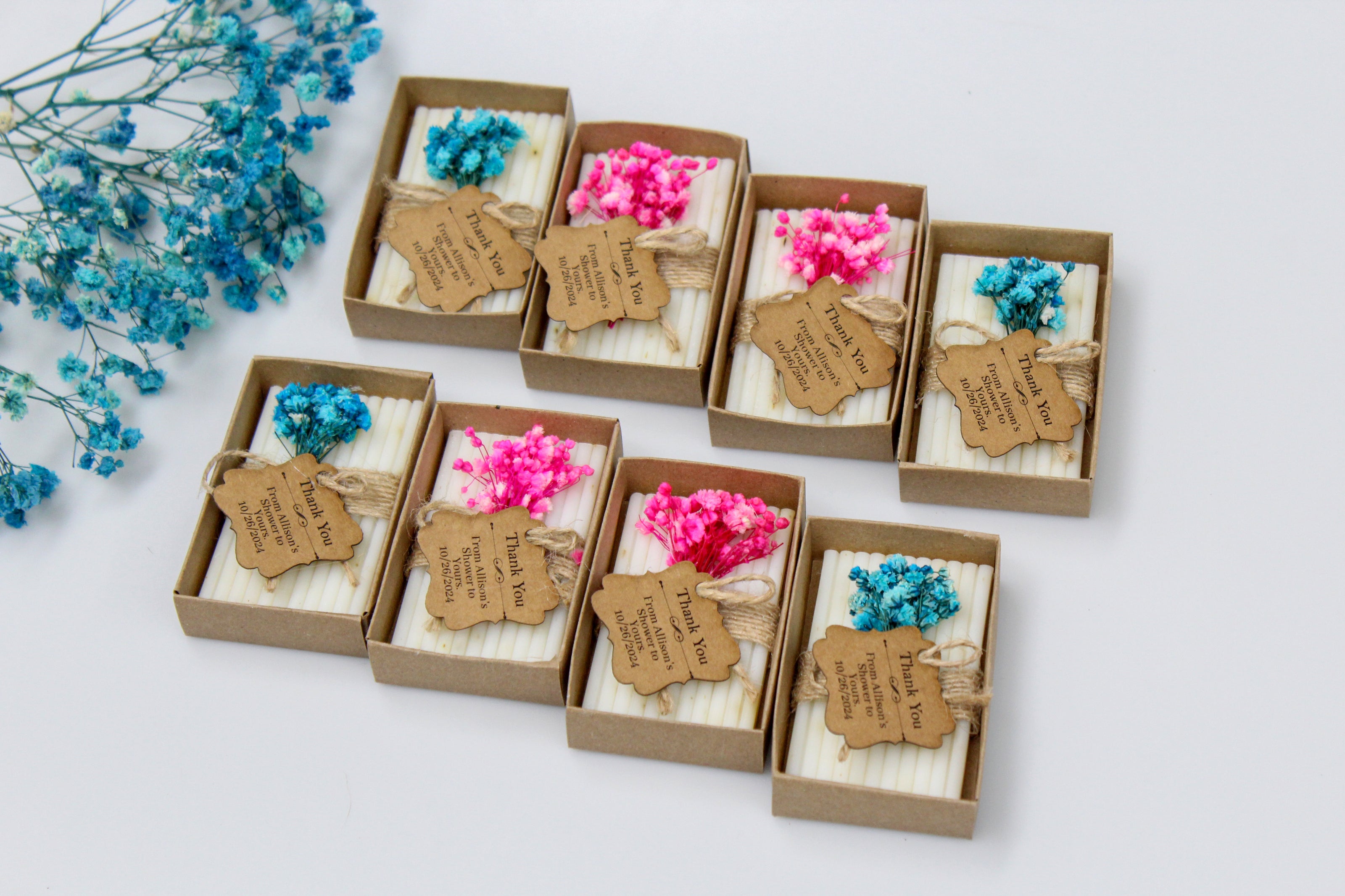 Personalized baby shower favors displayed in kraft paper boxes, featuring customizable thank you tags. Example tags read 'Thank You from Allison's Shower to Yours 10/26/2024'. Each favor is adorned with dried pink and blue flowers and natural twine, making them perfect for adding a personalized touch to baby shower celebrations.