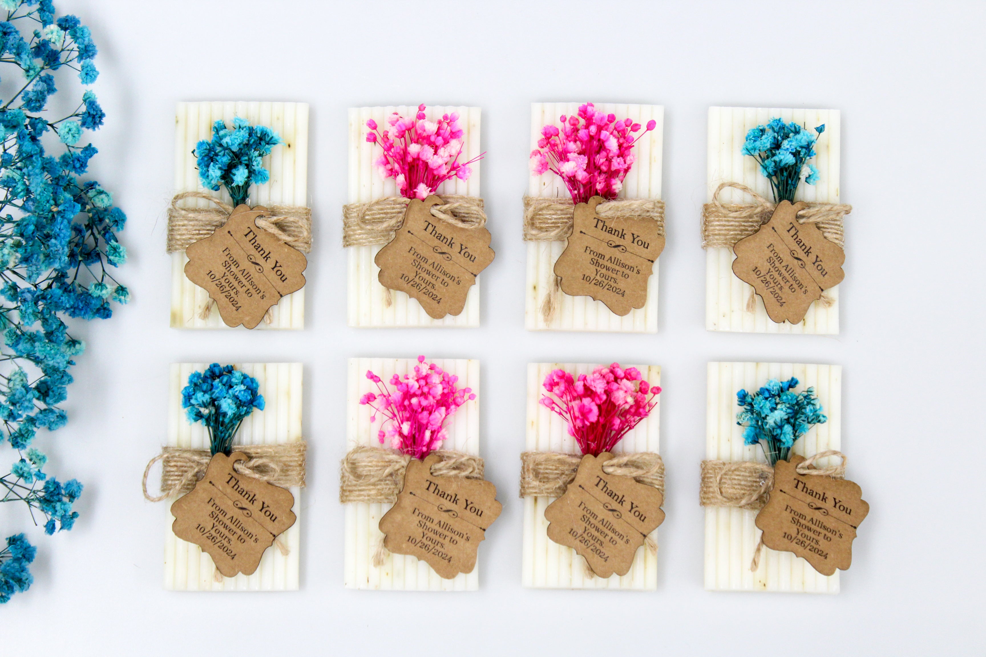 Assortment of personalized baby shower favors with custom thank you tags. Each favor displays a message such as 'Thank You from Allison's Shower to Yours 10/26/2024', featuring dried pink and blue flowers tied with natural twine. These eco-friendly favors are ideal for baby showers, adding a unique and personal touch to your celebration