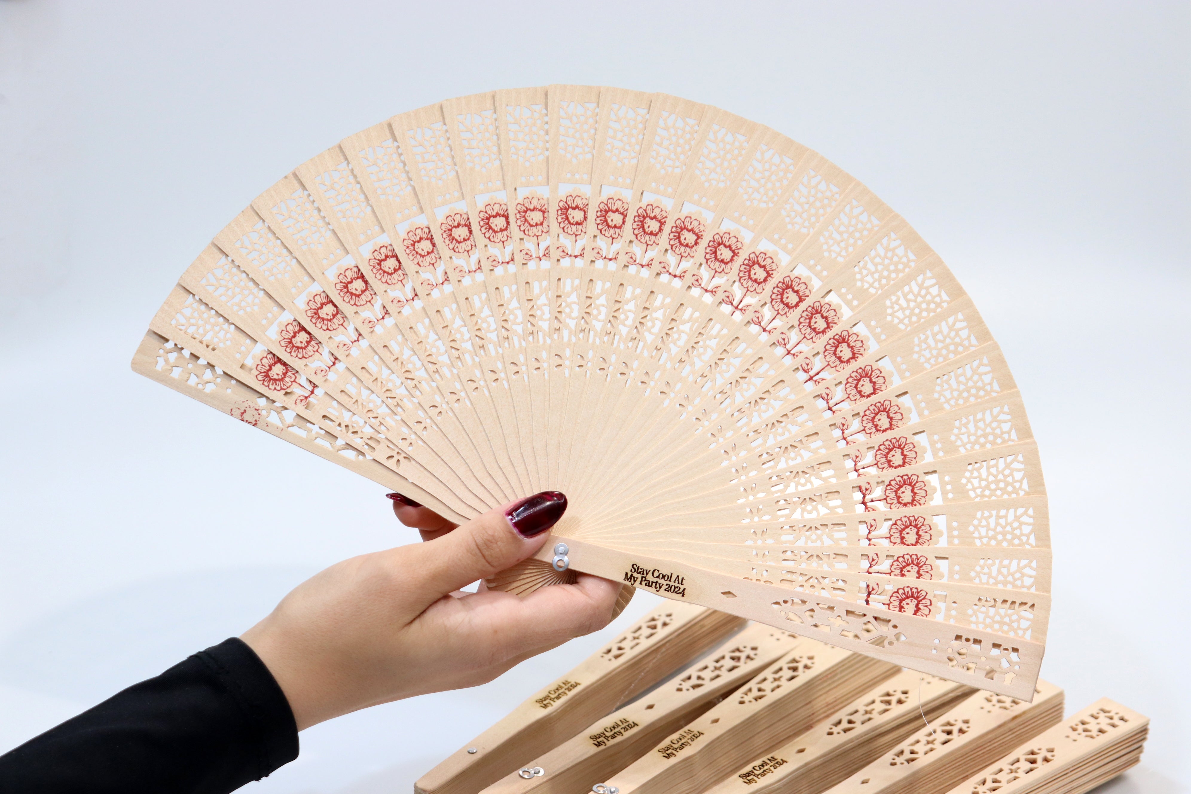 Hand holding a personalized rustic wooden hand fan with intricate lace patterns and red floral designs, ideal for party favors, featuring custom engraving 'Stay Cool At My Party 2024'.