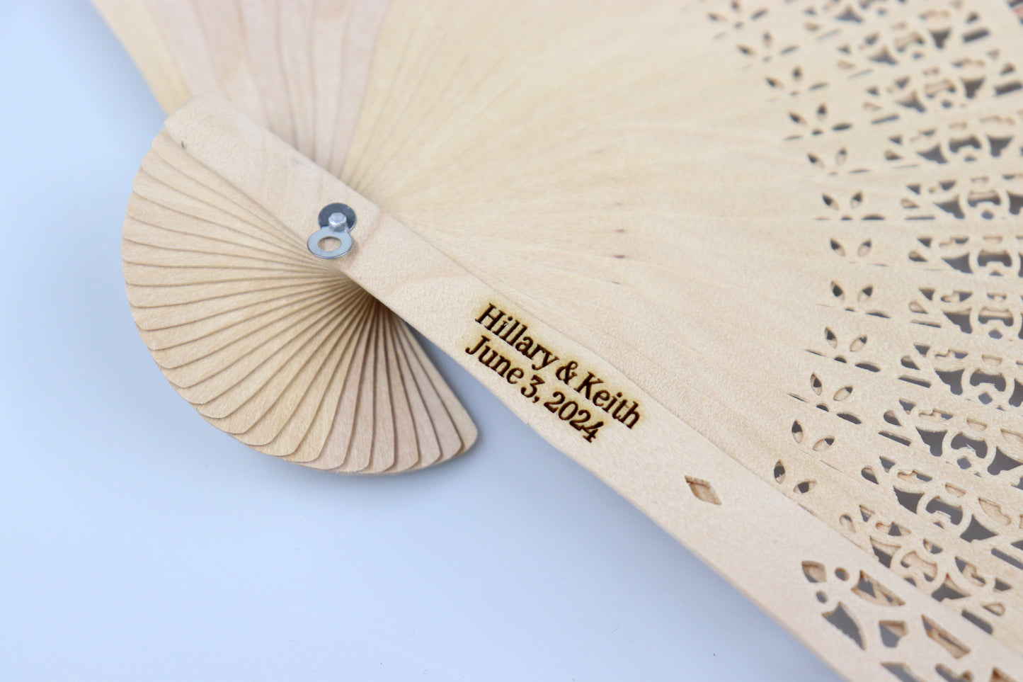 Close-up of a personalized rustic wooden hand fan with intricate lace patterns, engraved with 'Hilary & Keith June 3, 2024', perfect for wedding favors.