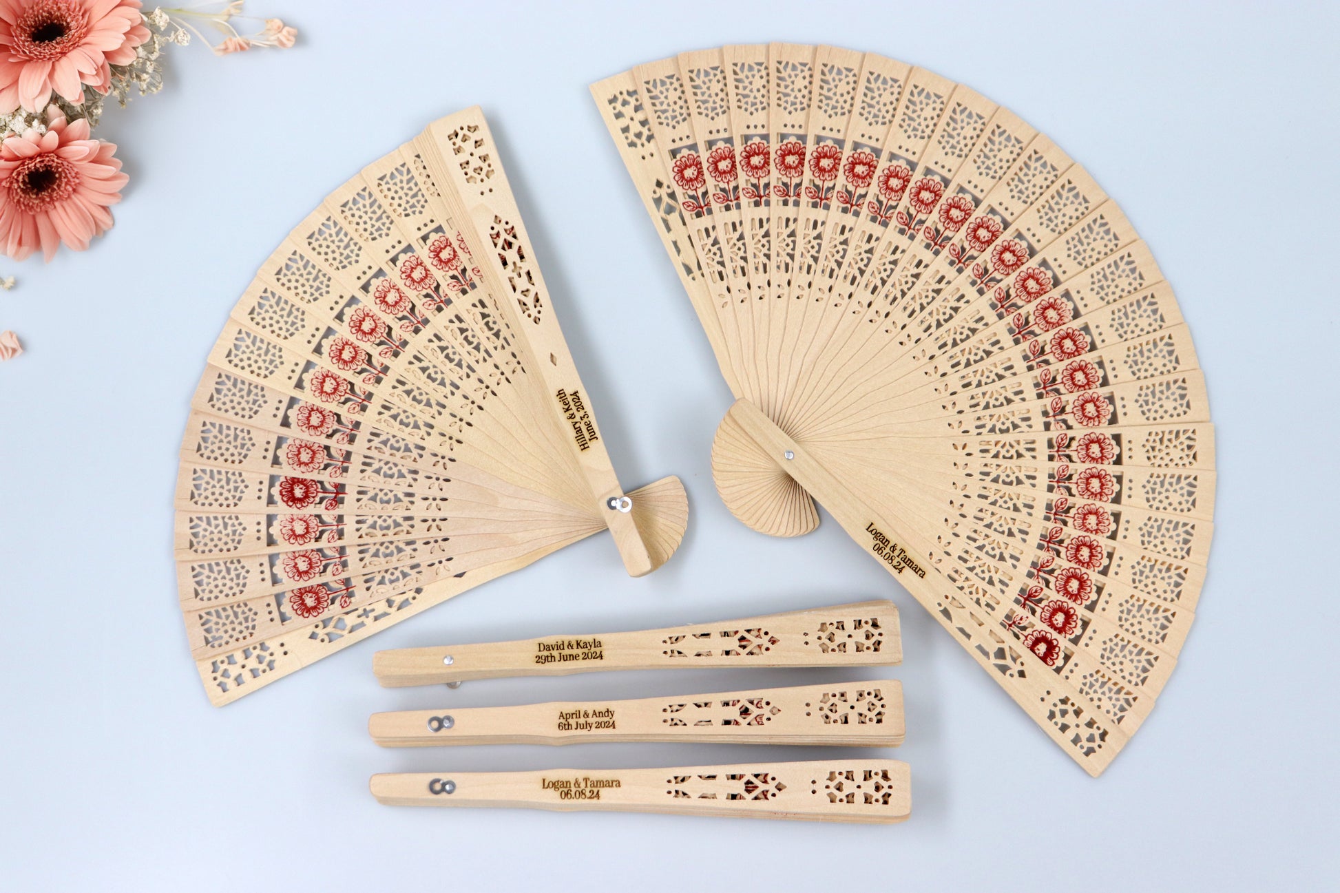 Set of personalized rustic wooden hand fans with intricate lace patterns and red floral designs, engraved with wedding dates and names, perfect for wedding favors, displayed with peach flowers on a light blue background.