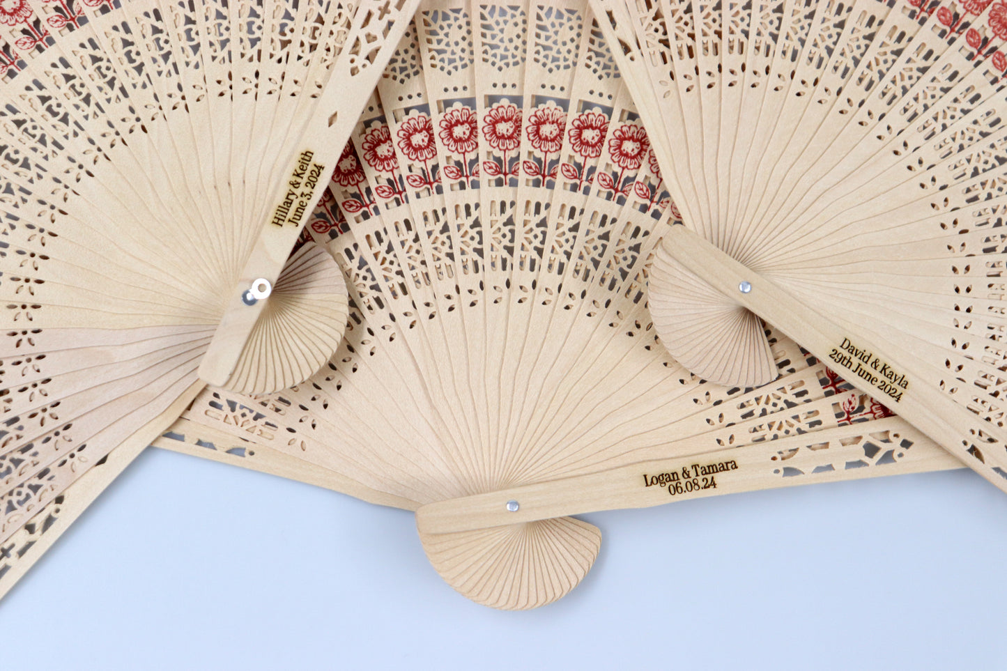 Collection of personalized rustic wooden hand fans with intricate lace patterns and red floral designs, engraved with wedding dates and names, ideal for wedding favors.