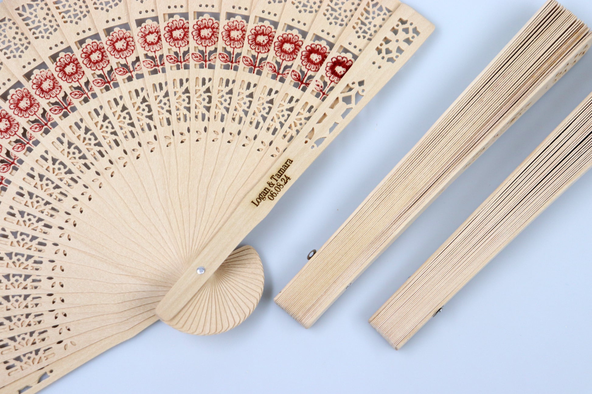 Rustic wooden hand fan with intricate lace patterns and red floral designs, personalized with 'Logan & Tamara 06.08.24', perfect for wedding favors, displayed next to its closed counterpart.