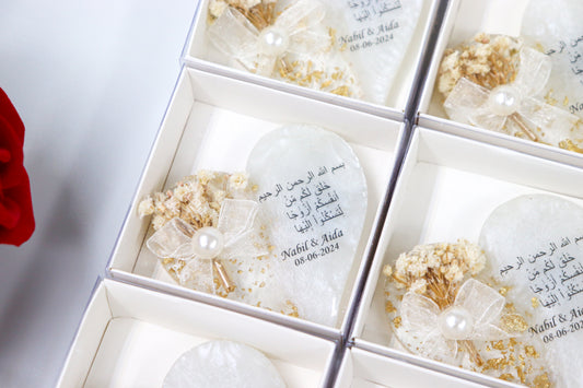 Close-up of elegant wedding favors in white boxes featuring heart-shaped personalized gifts with Arabic inscriptions and dried flower arrangements, perfect for special occasions, with red roses in the background.