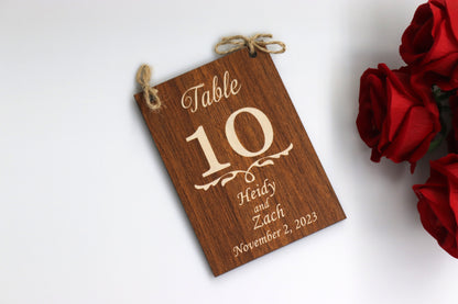 A personalized wooden table number sign for a wedding, displaying 'Table 10' with the names 'Heidy and Zach' and the date 'November 2, 2023.' The sign features a dark wood finish and is laid flat on a white surface, with red roses in the background.