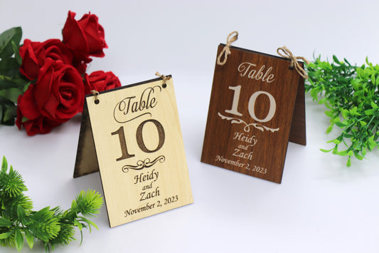 Example of personalized wooden table numbers for wedding receptions. Two wooden signs with 'Table 10' and example names 'Heidy and Zach' with the date 'November 2, 2023,' displayed in light and dark wood finishes. Decorated with rope for easy hanging, with red roses and greenery in the background.