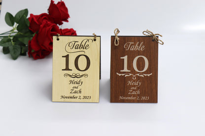 Two examples of personalized wooden table number signs for a wedding, displaying 'Table 10' with the names 'Heidy and Zach' and the date 'November 2, 2023.' The sign on the left features a light wood finish, while the sign on the right features a dark wood finish. Both signs are adorned with twine for hanging, with red roses in the background.