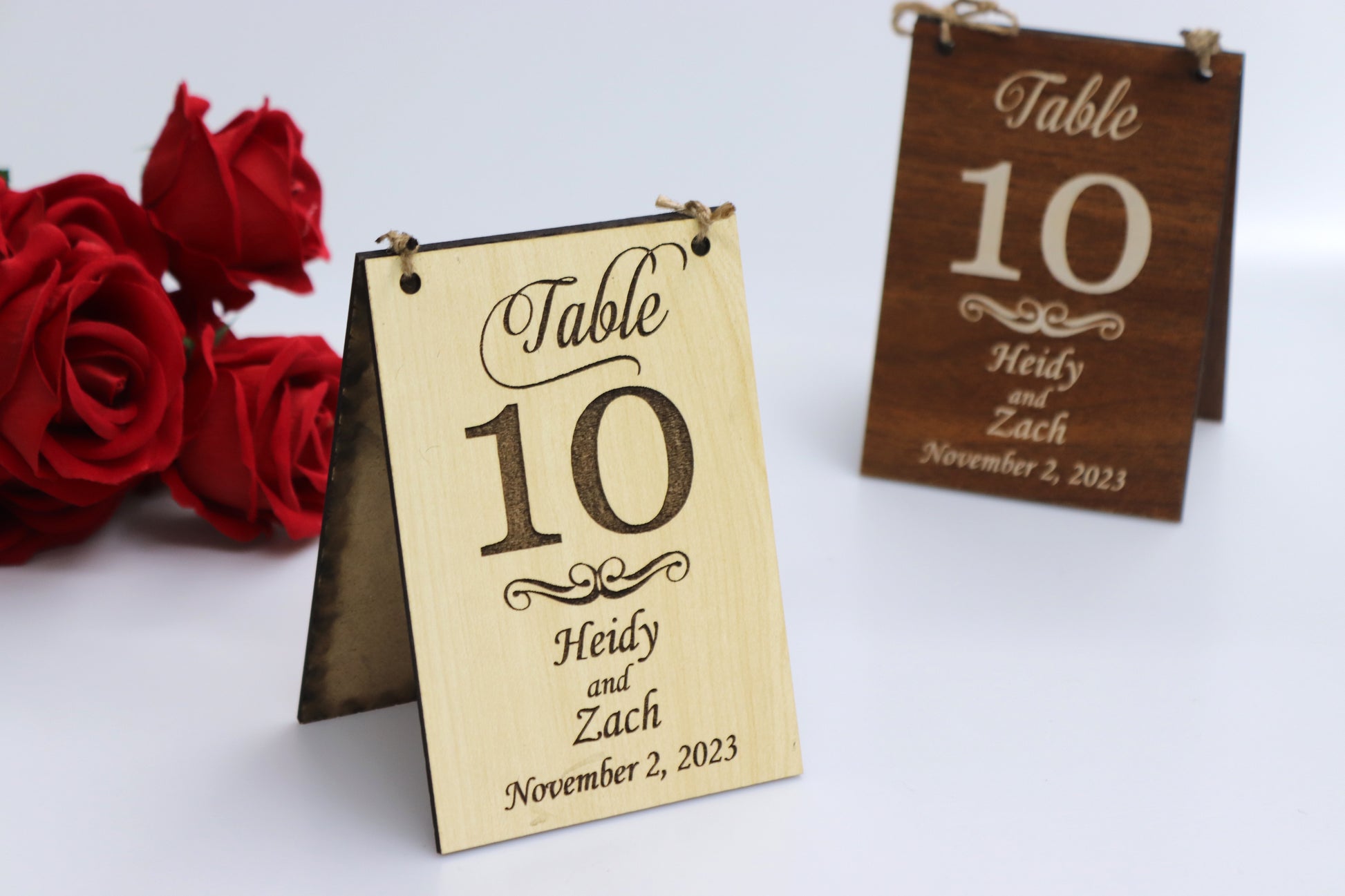 Personalized wooden wedding table signs in light and dark wood with table number, names, and date example, set against a white background with red roses. Perfect for rustic or vintage-themed weddings