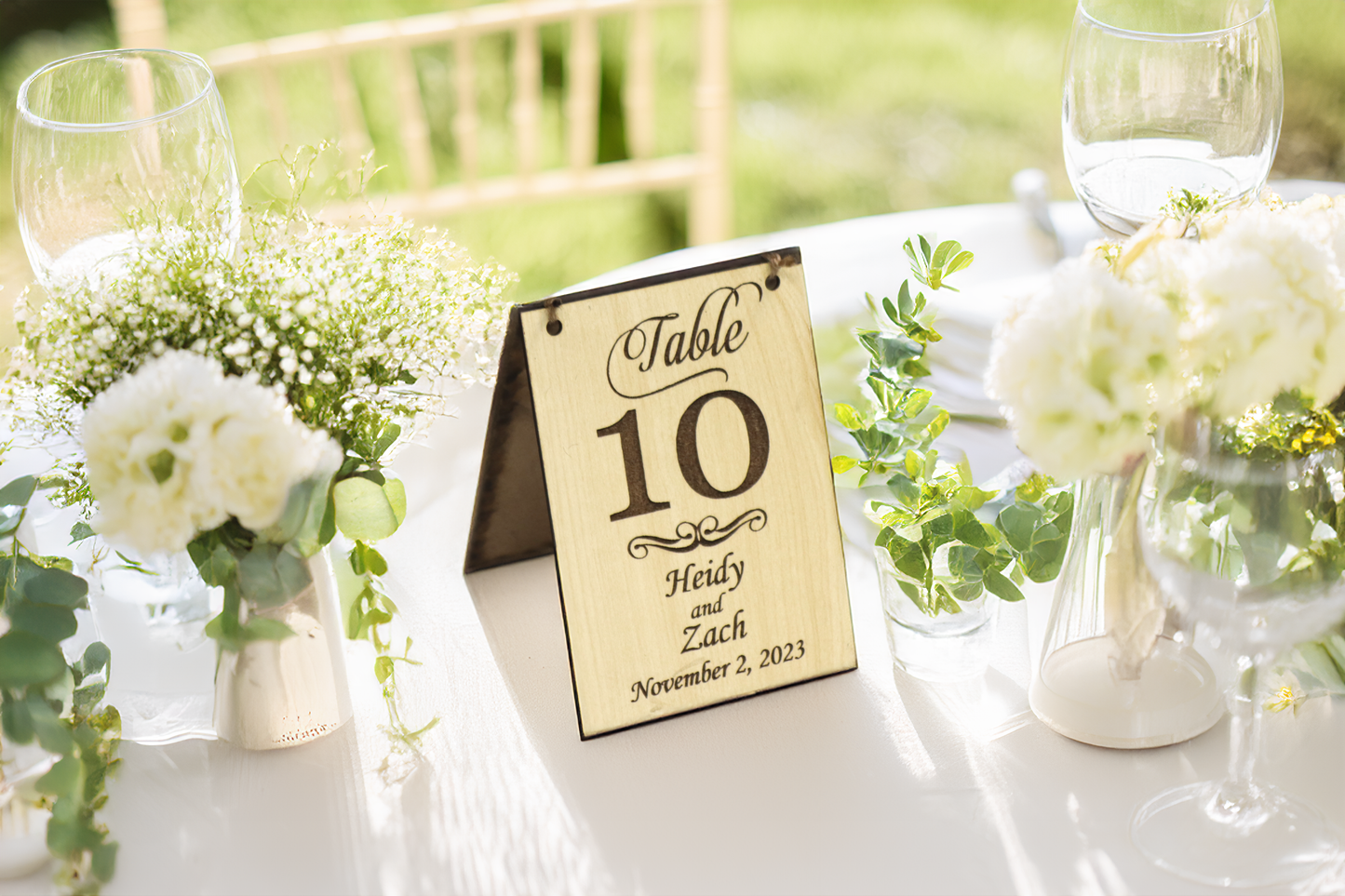 Example of a personalized wooden table number sign for a wedding reception. The sign displays 'Table 10' with example names 'Heidy and Zach' and the date 'November 2, 2023,' on a light wood finish. It is placed on a beautifully decorated table with white flowers, greenery, and elegant glassware, set in an outdoor setting.