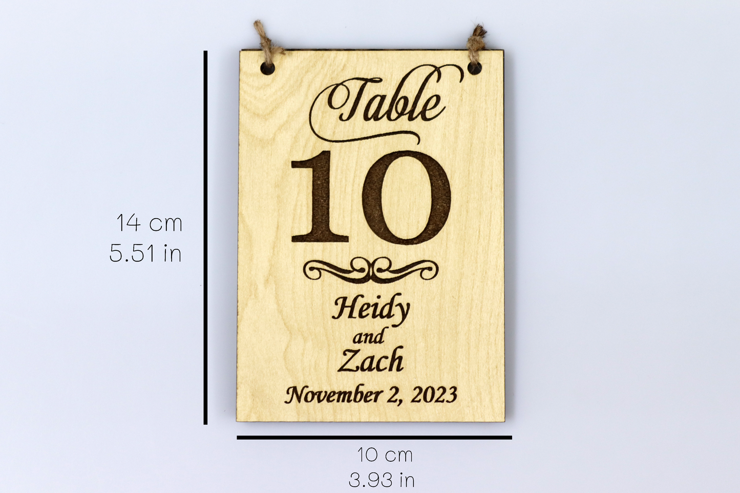 Example of a personalized wooden table number sign for a wedding, displaying 'Table 10' with the names 'Heidy and Zach' and the date 'November 2, 2023.' The sign's dimensions are indicated as 14 cm (5.51 in) in height and 10 cm (3.93 in) in width.