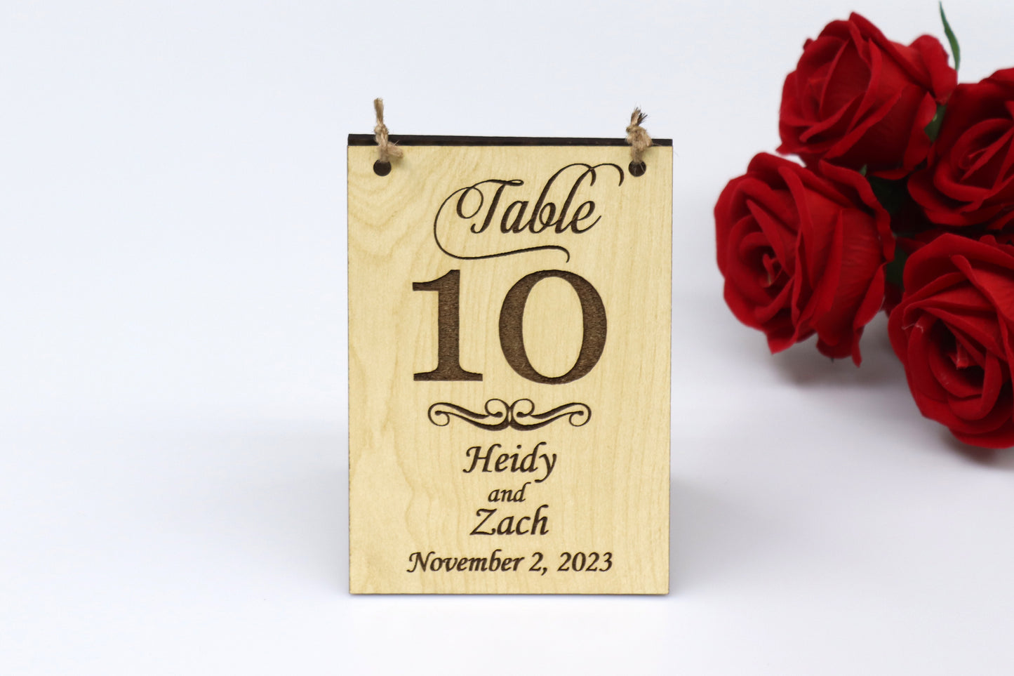 Rustic personalized wooden wedding table sign with table number 10, names Heidy and Zach, and date November 2, 2023, example displayed on a light wood background with red roses on the side. Ideal for vintage and rustic-themed weddings