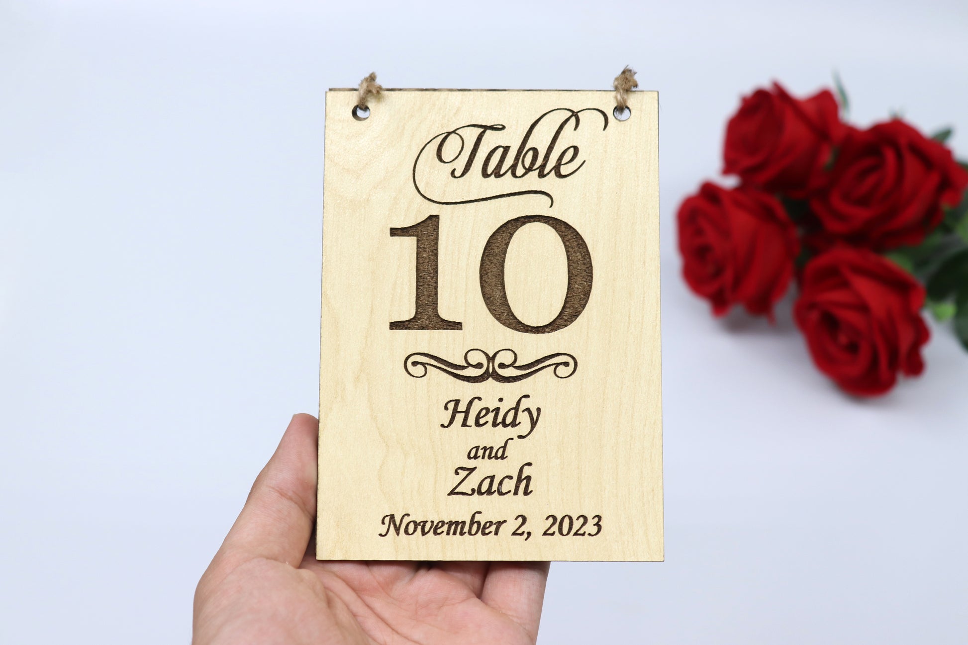 A personalized wooden table number sign for a wedding, displaying 'Table 10' with the names 'Heidy and Zach' and the date 'November 2, 2023.' The sign features a light wood finish and is being held in hand, with red roses in the background.