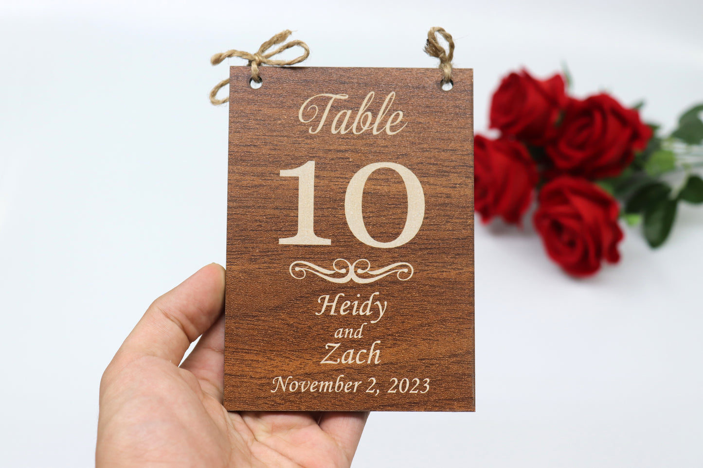 Example of a personalized wooden table number sign for a wedding, displaying 'Table 10' with the names 'Heidy and Zach' and the date 'November 2, 2023.' The sign features a dark wood finish and is shown being held by a hand, with red roses in the background.