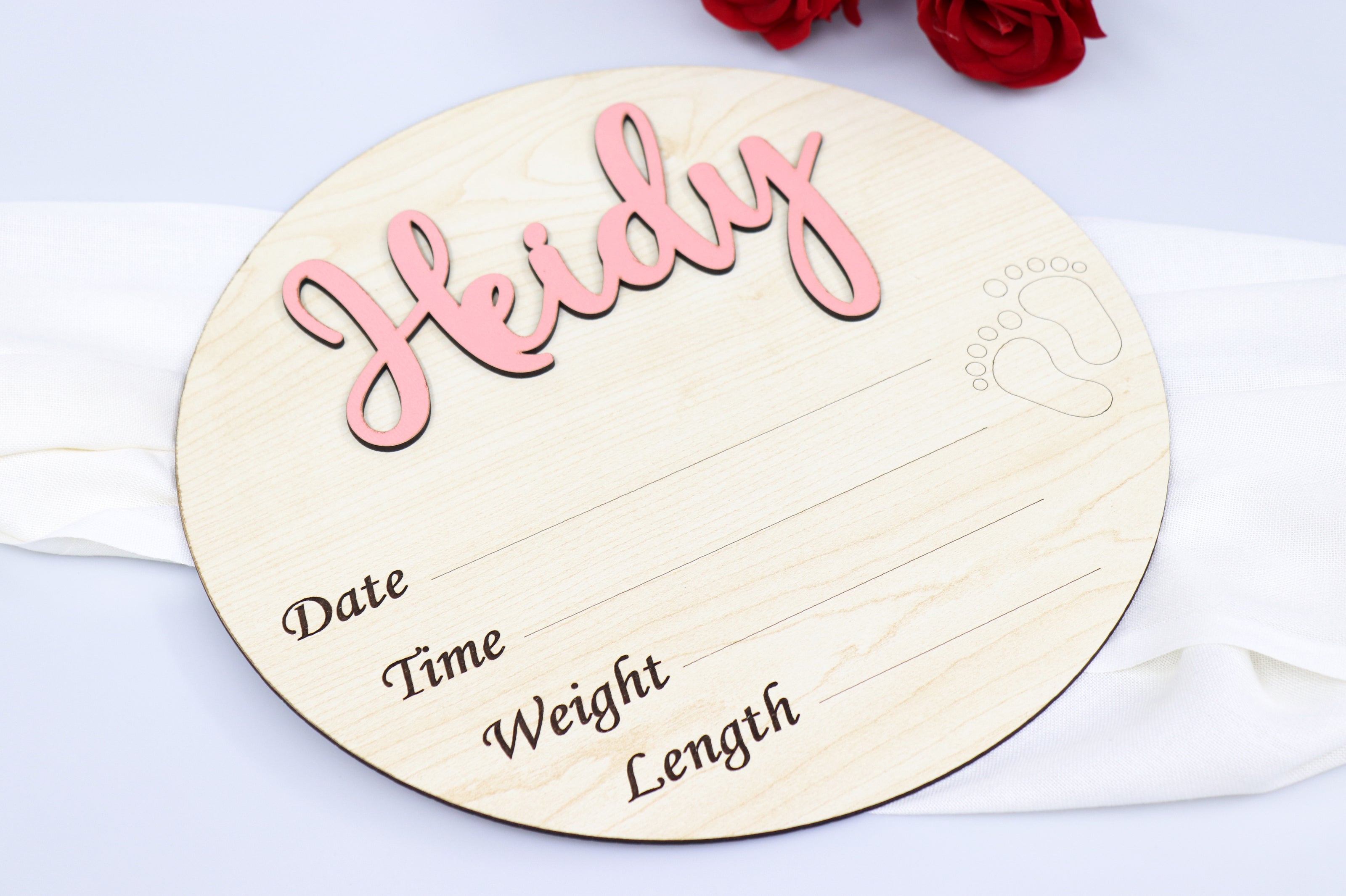 Personalized wooden birth announcement sign with the name 'Heidy' in pink script. The round sign includes customizable fields for date, time, weight, and length, along with an engraved footprint design. Perfect for a memorable keepsake or a thoughtful baby shower gift.