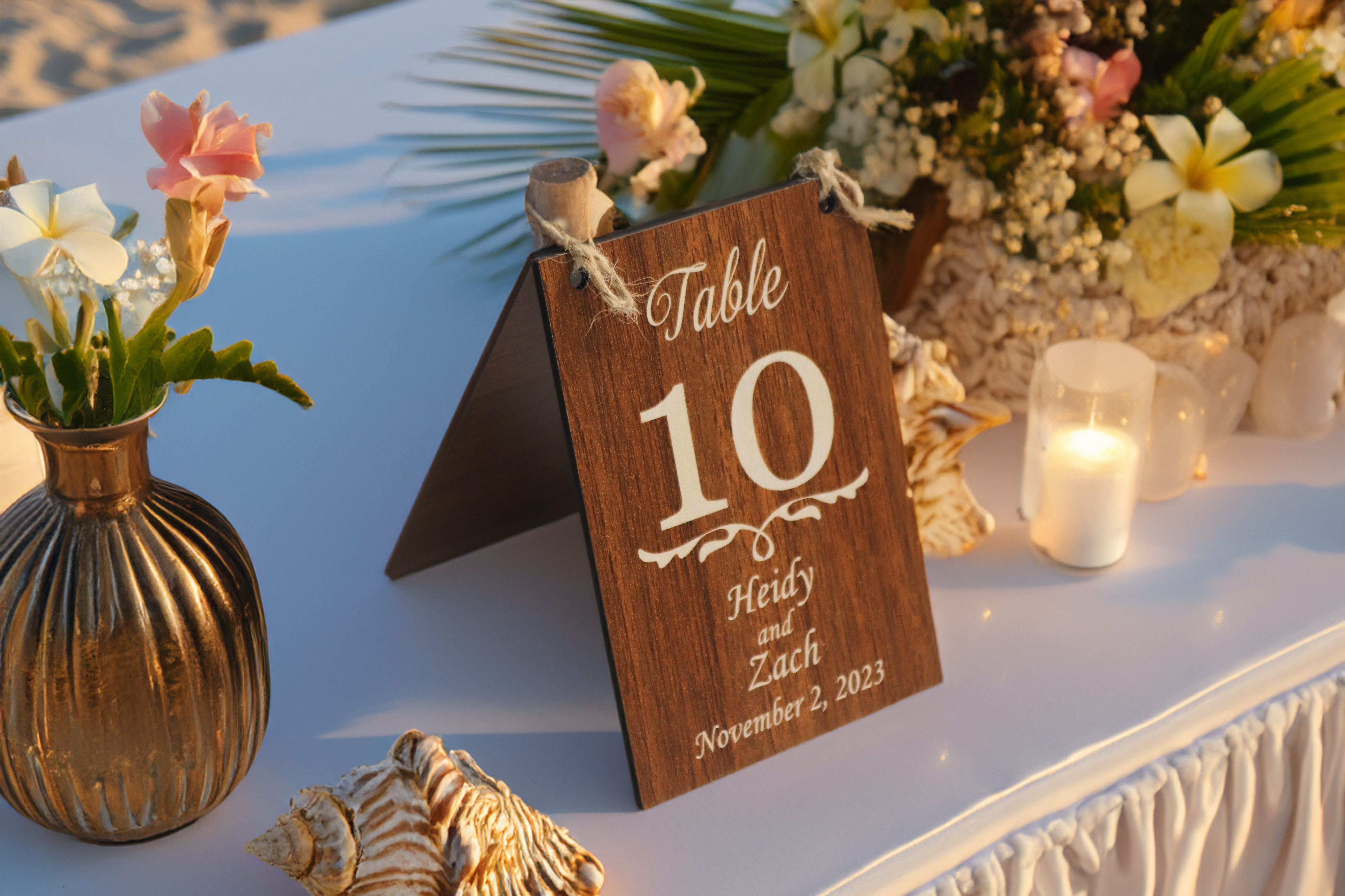 A personalized wooden table number sign for a wedding, displaying 'Table 10' with the names 'Heidy and Zach' and the date 'November 2, 2023.' The sign features a dark wood finish and is set on a white table adorned with flowers, seashells, and a lit candle, creating an elegant beachside wedding ambiance