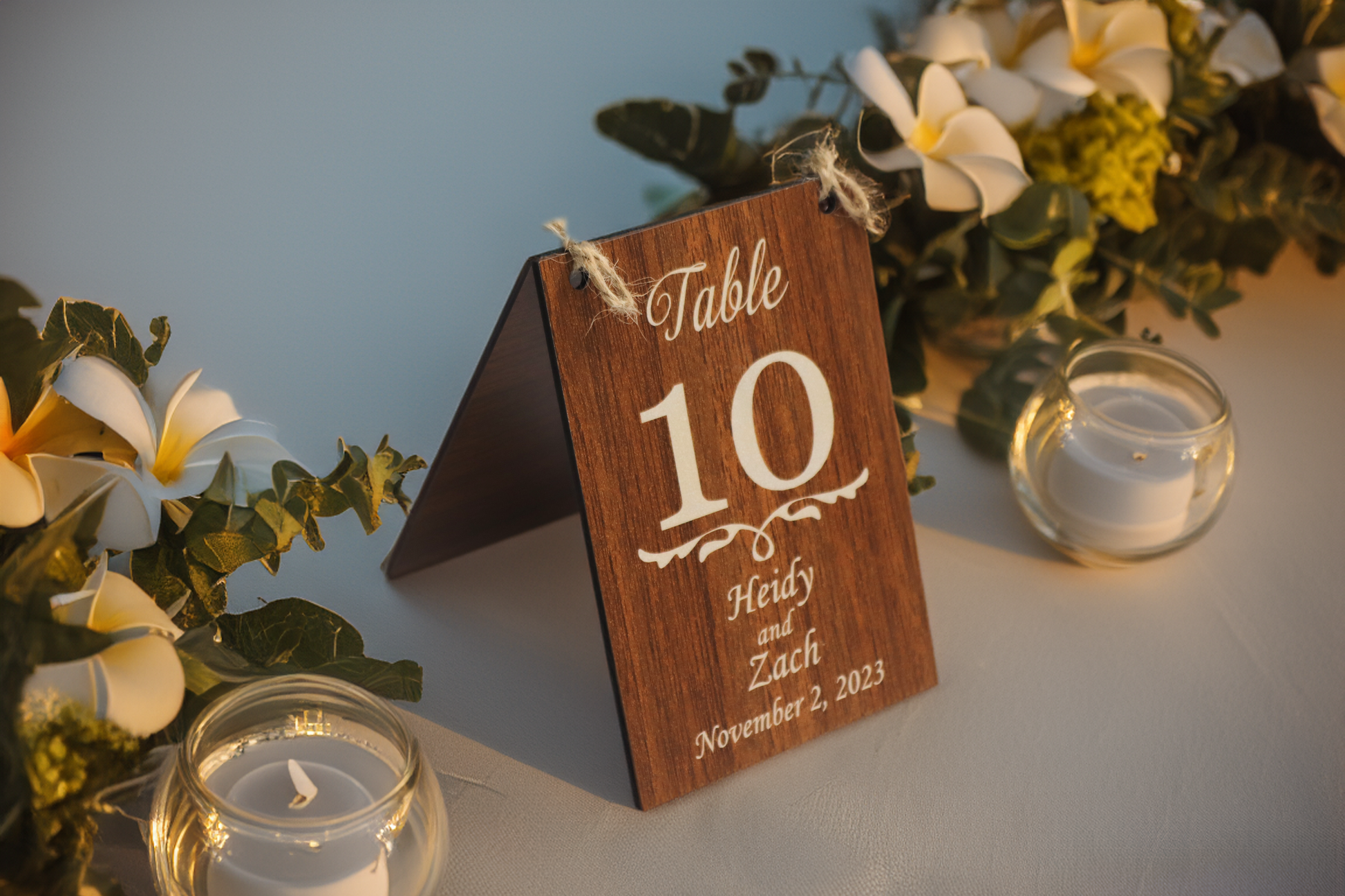 Example of a personalized wooden table number sign for a wedding reception. The sign displays 'Table 10' with example names 'Heidy and Zach' and the date 'November 2, 2023,' on a dark wood finish. It is decorated with rope for easy hanging and placed among white flowers and candles in glass holders.