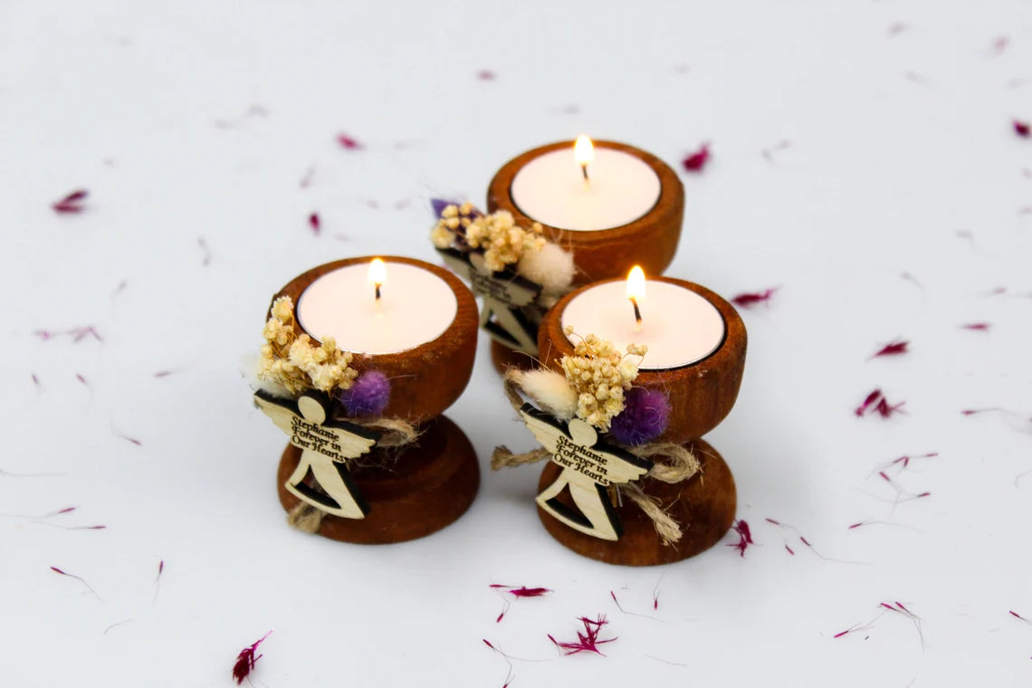 Set of three personalized handcrafted funeral favor candles. Each wooden candle holder is adorned with dried flowers and a small angel charm engraved with 'Stephanie Forever in Our Hearts.' The candles are lit, symbolizing remembrance and tribute to the loved one.