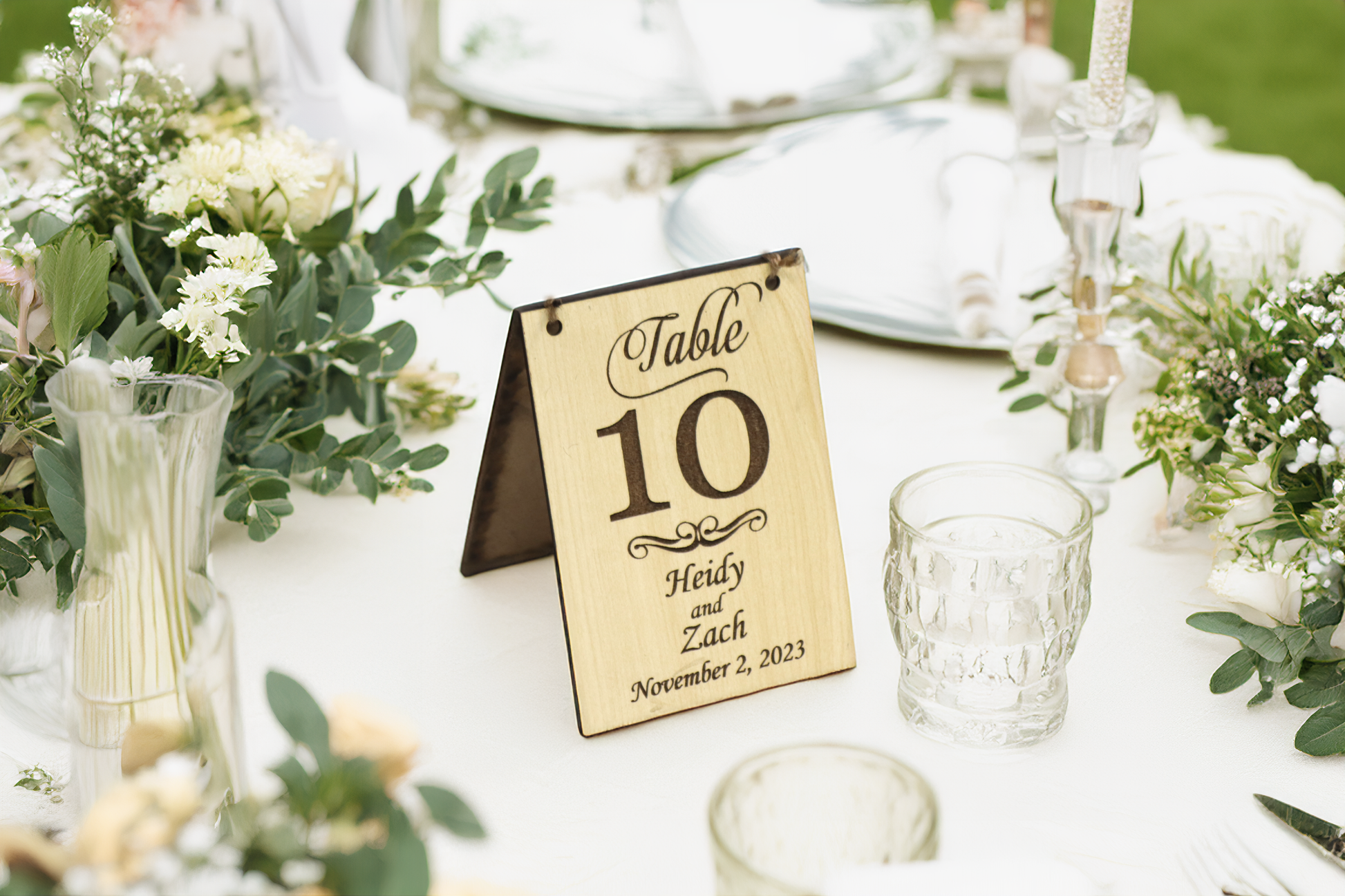 A personalized wooden table number sign for a wedding, displaying 'Table 10' with the names 'Heidy and Zach' and the date 'November 2, 2023.' The sign features a light wood finish and is elegantly placed on a table set with glassware, floral arrangements, and greenery.